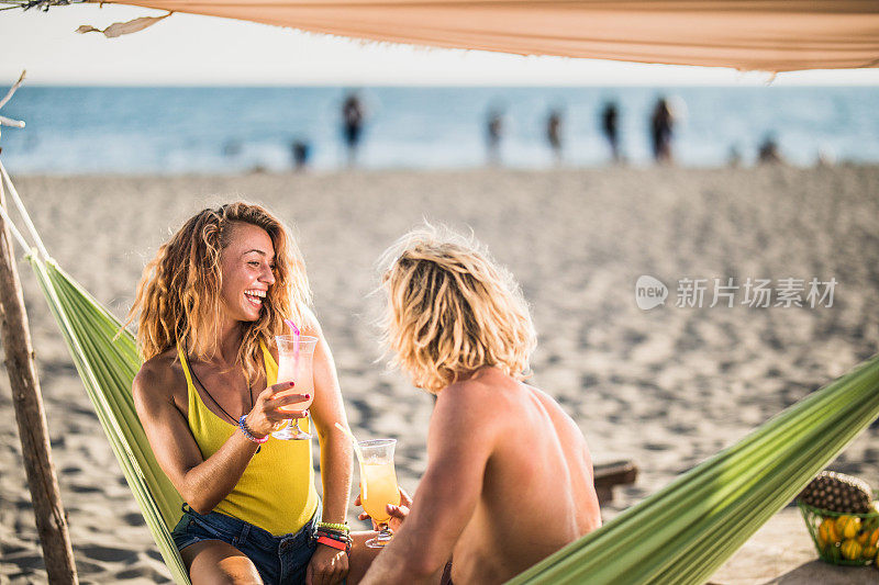 年轻幸福的夫妇在海滩上的吊床上喝着鸡尾酒聊天。