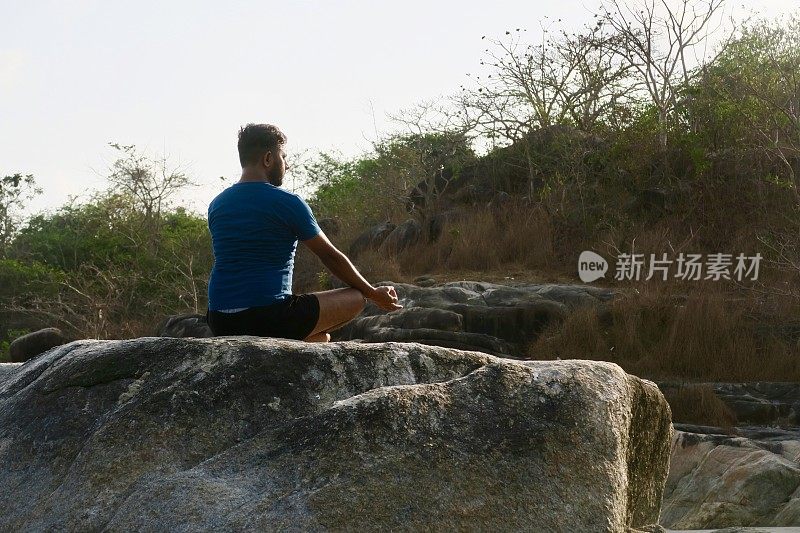 印度男子练习莲花姿势(莲花瑜伽)瑜伽位置在岩石上，后视图，水的边缘，帕洛伦海滩，果阿，印度