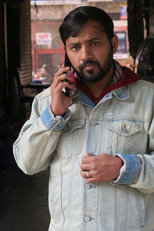 英俊的印度男子用手机在户外打电话叫出租车，印度，年轻的印度男子用手机说话交流，城市背景，好看的家伙穿着连帽衫牛仔夹克，走路谈话集中