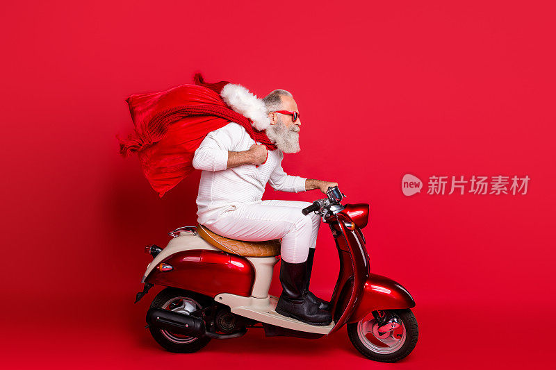 侧面的漂亮的胡子灰白头发的圣尼古拉斯骑电机送携带可取的购买的东西12月冬天孤立在明亮鲜艳的亮红色背景
