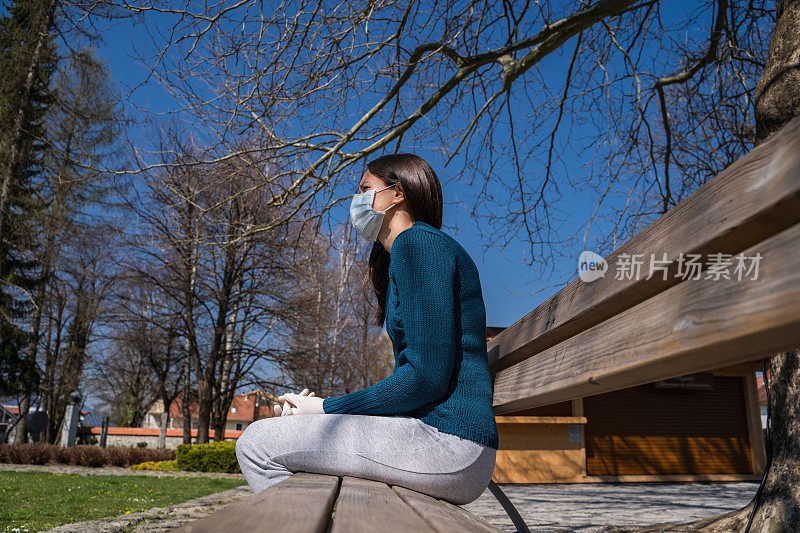 侧面的年轻妇女在外科口罩坐在长椅上在公园享受阳光明媚的天气-股票照片