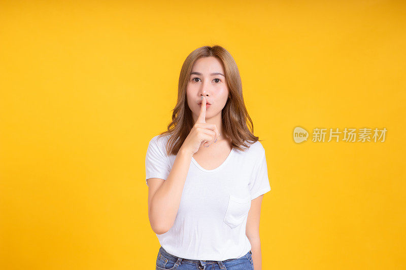 肖像年轻的亚洲女子手指在嘴唇嘘嘘安静的象征手势沉默反对在白色t恤，黄色背景孤立的工作室拍摄和复制空间。
