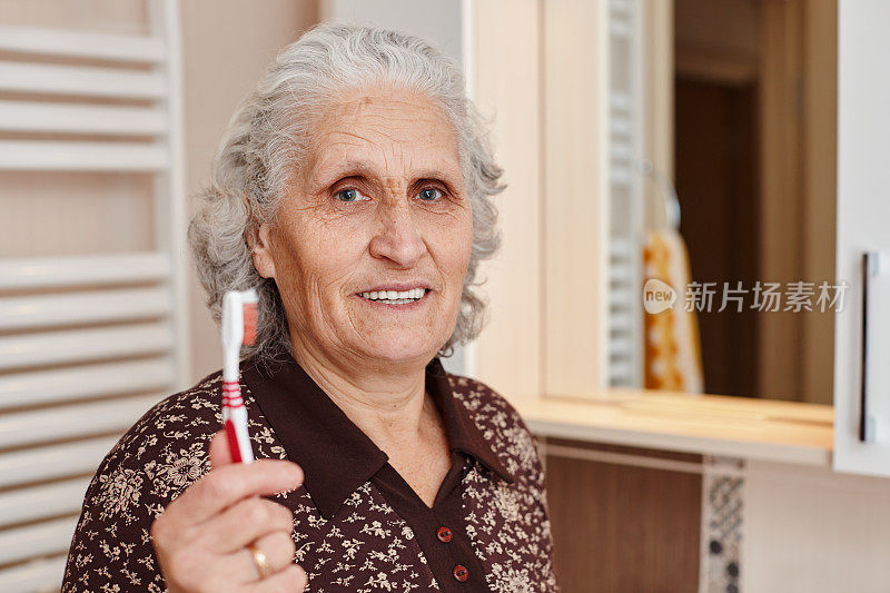 美丽的老年妇女与灰色头发拿着一个牙刷在手里，而看着镜头与一个巨大的牙齿微笑在浴室的镜子和水槽