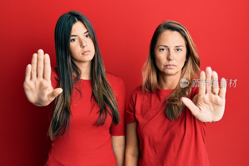 西班牙家庭的母亲和女儿穿着休闲衣服在红色的背景做停止与手掌唱歌。警告表情与否定和严肃的手势在脸上。