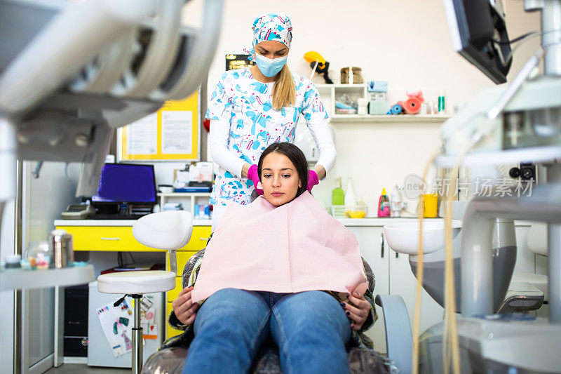 一位年轻女子坐在牙科治疗椅上，她的牙医正在系一次性牙围兜