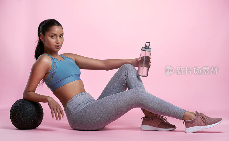 一个迷人的和运动的年轻女子与一个药球和水瓶在工作室在粉红色的背景摆姿势的全长肖像