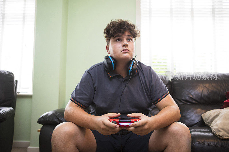 一个十几岁的男孩在玩电子游戏，脖子上戴着耳机。他穿着灰色的t恤和短裤。