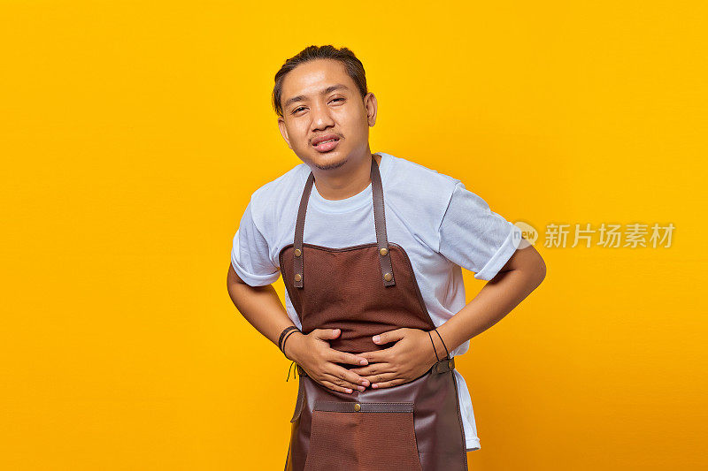 亚洲年轻男子的肖像戴着围裙，双手放在胃由于恶心，痛苦的疾病感觉不适