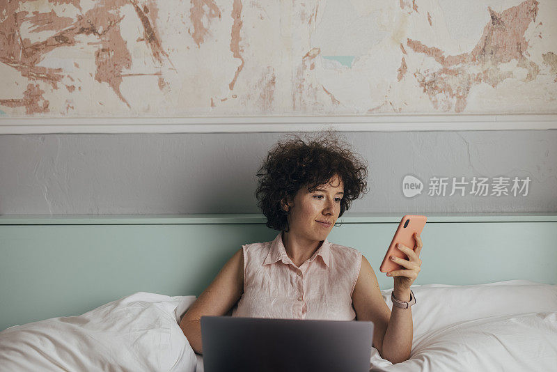 一名年轻的白人女性在床上看着手机，膝上放着笔记本电脑