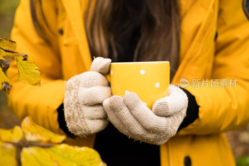 秋天的森林里，穿着黄色皮大衣的女人手里拿着黄色的茶杯。