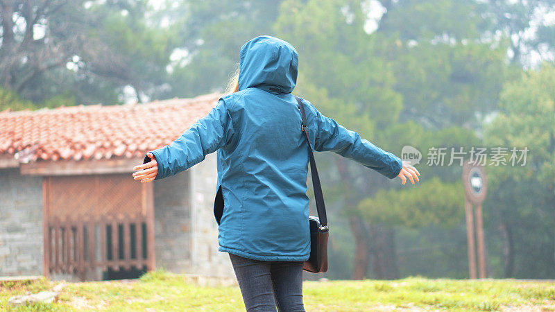 女性的后视图。她在大自然中张开双臂散步。她享受着自由和宁静。季节是秋天。她穿着一件蓝色的雨衣和深蓝色的牛仔裤。