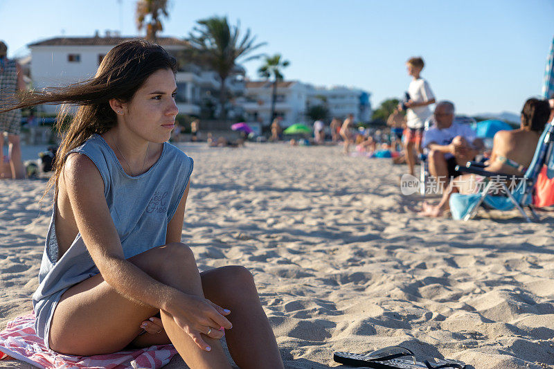 下午晚些时候，西班牙马略卡岛海滩上，一位年轻女子坐在沙滩上的毛巾上。