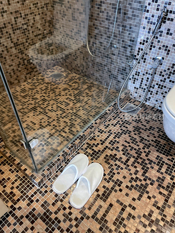 除了浴室玻璃淋浴间外，白色马赛克瓷砖地板上的白色酒店拖鞋的图像，白色陶瓷马桶，手淋浴坐浴盆头附在墙上，重点放在前景