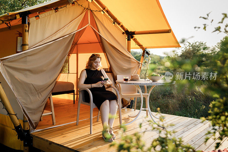 快乐的女人自由职业者使用电话和喝鸡尾酒电话，而放松在舒适的豪华帐篷在阳光明媚的日子。豪华户外露营帐篷，适合夏季度假度假。生活方式的概念