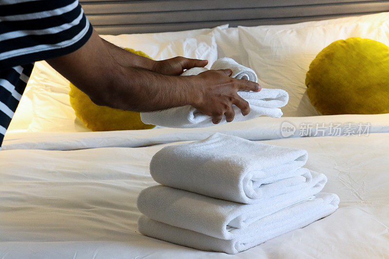 特写图像:一个认不出的人把一堆白色酒店毛巾放在白色羽绒被双人床上，折叠的浴巾和手巾，圆形的黄色丝绒靠垫靠在白色枕头上，靠在有纹理的棒状床头板上