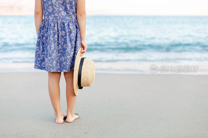小女孩在海滩上的暑假。穿蓝裙子戴草帽的可爱小孩是游客。儿童假期，旅游，温柔，女性化的概念。孩子在海边户外散步。