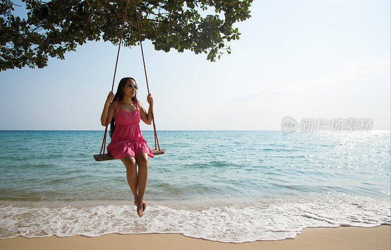 美丽的亚洲女人放松在热带海滩上的秋千。