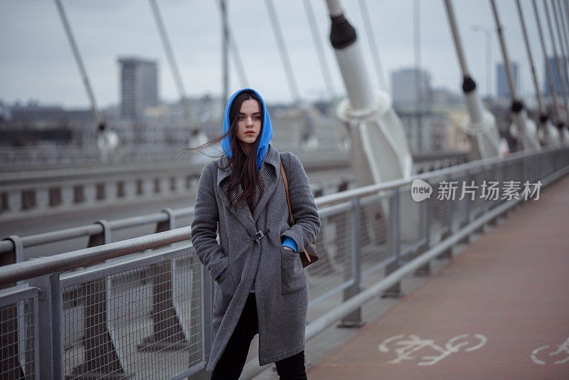 穿着大衣的年轻女子，在刮风的天气走在城市的桥上。都市秋冬风格