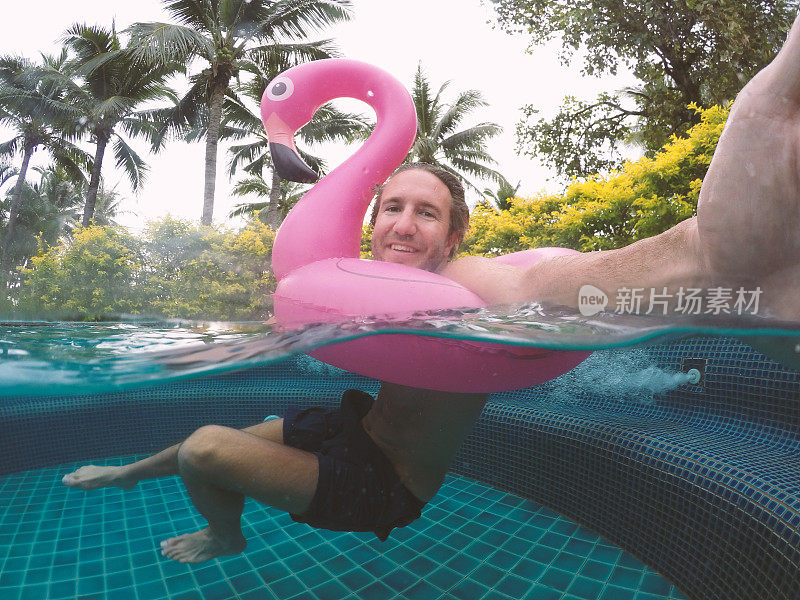 在度假村游泳池里玩充气火烈鸟的时候，一个快乐的男人在水下自拍。热带趣味度假概念