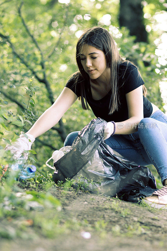 女志愿者穿着衣服在公园里捡垃圾和塑料垃圾。小女孩戴着手套，在户外把垃圾放进黑色的塑料袋里。