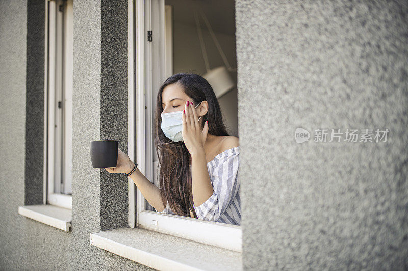 一名年轻的成年女子，因冠状病毒被隔离期间，靠向打开的窗户，向窗外望去，喝着咖啡，头痛，戴着医用口罩