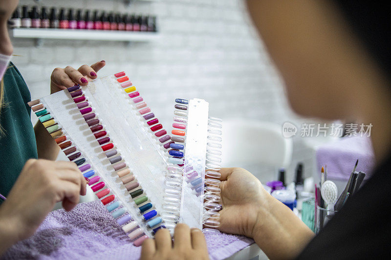 年轻女子看指甲颜色调色板-彩色的人工指甲在一个美甲店-一个年轻女子选择她的指甲的颜色，而美容师帮助她选择-指向指甲油的颜色