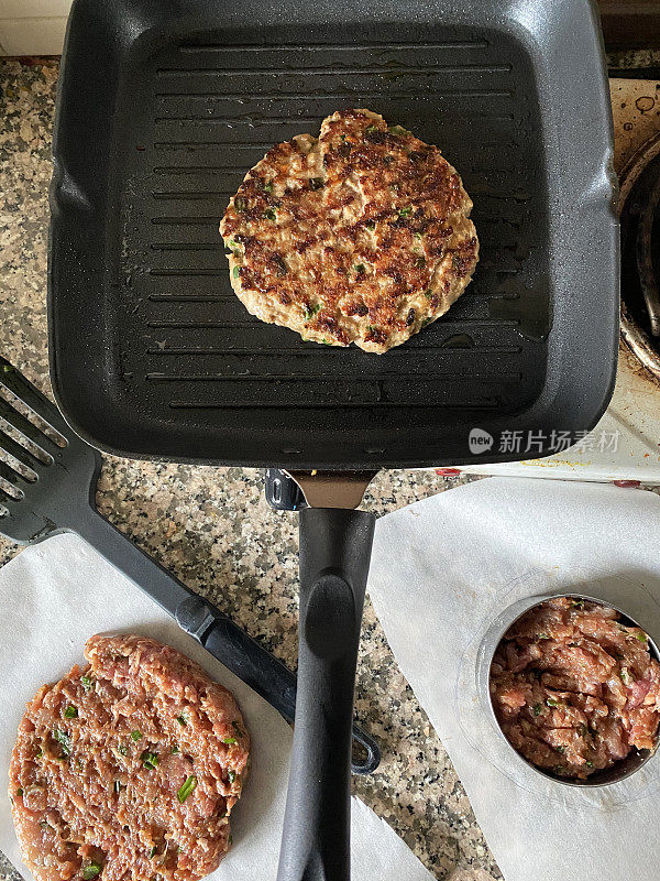 不粘锅里装着自制牛肉汉堡肉饼，由碎肉和洋葱在煎锅里煎成，旁边是生肉饼，放在防油纸上，厨房场景，俯瞰