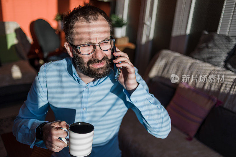 一个年轻的有前途的商人正在休息和喝咖啡，同时使用一个模型手机，是在窗户附近-在家里工作。