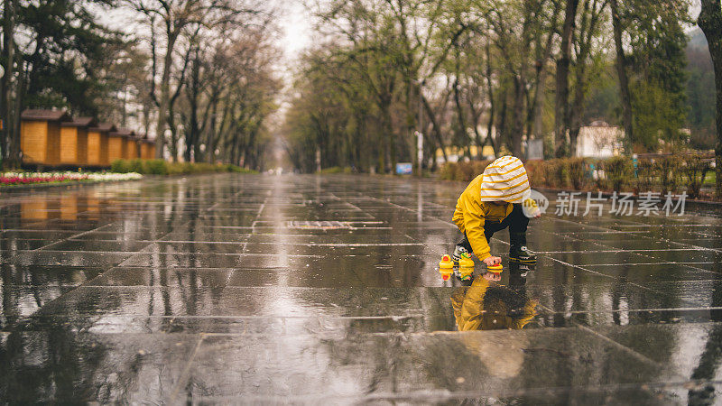 可爱活泼的小男孩穿着明黄色雨衣和胶靴在潮湿的春雨日街道上的小水坑里玩橡皮鸭。季节性天气步行概念