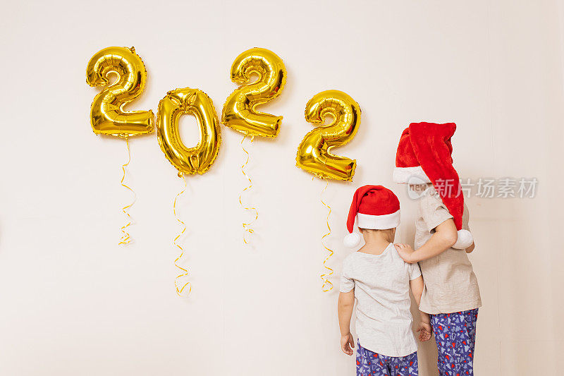 孩子们戴着红色的圣诞帽，在白色的墙上挂着金色的气球，享受着五彩纸屑。在家里庆祝2022年新年。圣诞节的概念