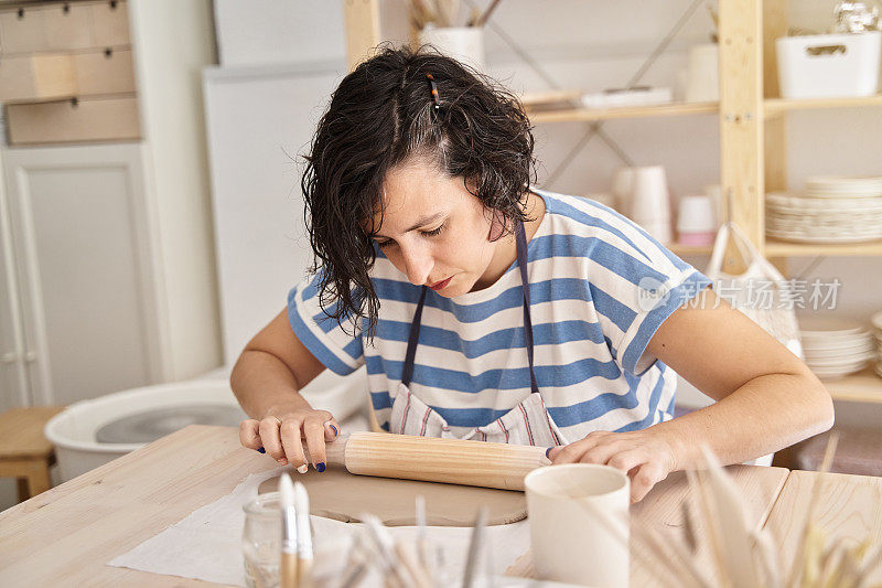 一名身穿蓝色条纹衬衫的妇女在陶器作坊里揉粘土。女人正在学习制作粘土