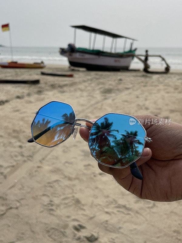一副八角形，金属框架，有色，镜面太阳镜的图像，由一个无法辨认的人拿着，反射出棕榈树，沙滩，色彩鲜艳的船在水边，海浪在海滩上破碎的海岸背景，重点在前景