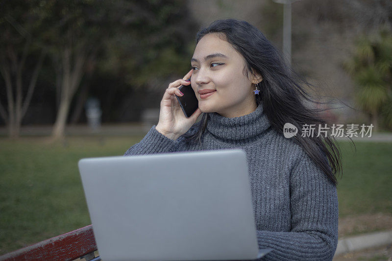 年轻的亚洲妇女在公园里一边用电脑一边打电话