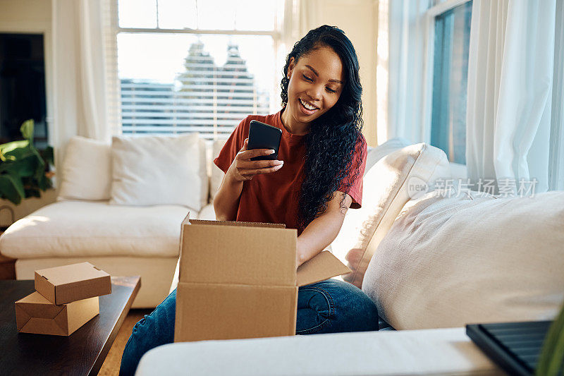 快乐的黑人妇女使用手机，同时打开家庭递送的包裹。