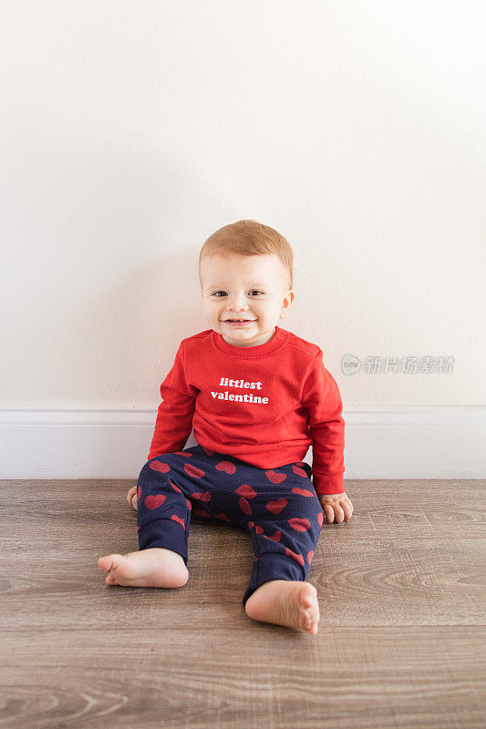 2023年2月的情人节，一个可爱而快乐的14个月大的小男孩笑着，光着脚，穿着写着“最小的情人节”的红色毛衣和印有红心的海军慢跑裤，站在木制硬木地板上