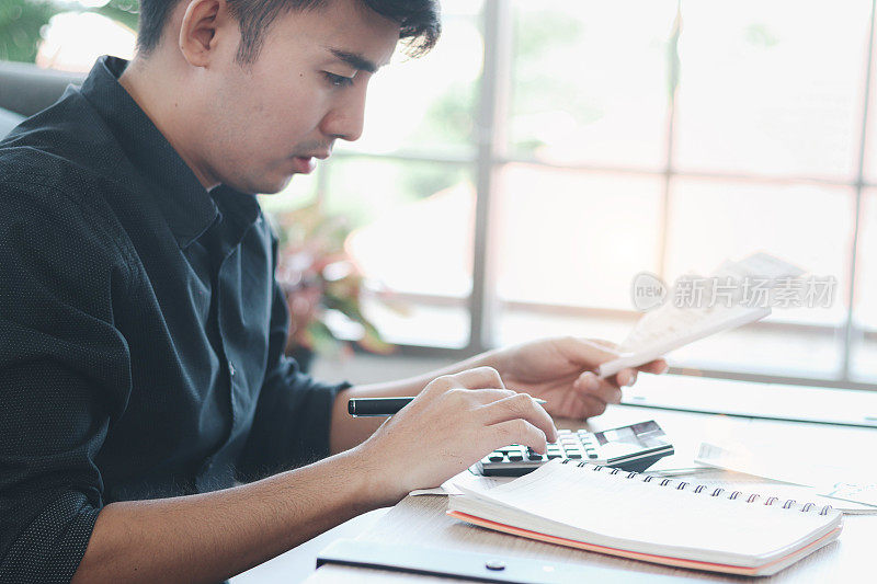 穿着黑色衬衫的年轻商人用计算器计算他的账单，思考他的财务信息和债务问题，想要减少开支和省钱。