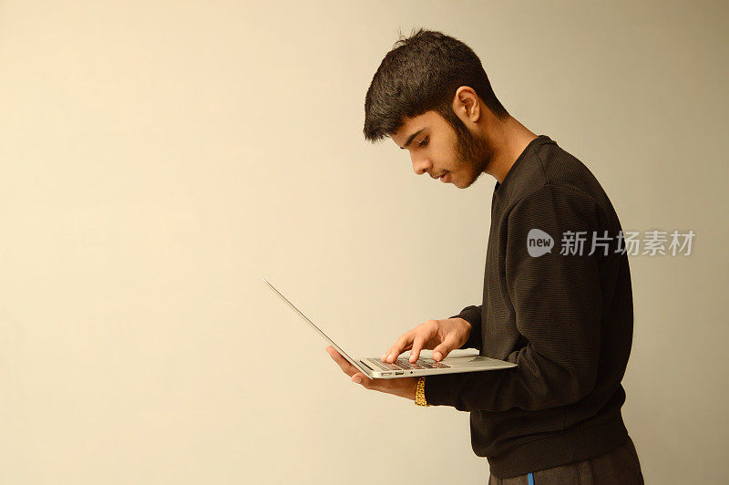 一个年轻的印度亚裔少年，留着胡子，手里拿着笔记本电脑，专注地盯着屏幕，一边站着一边忙着工作