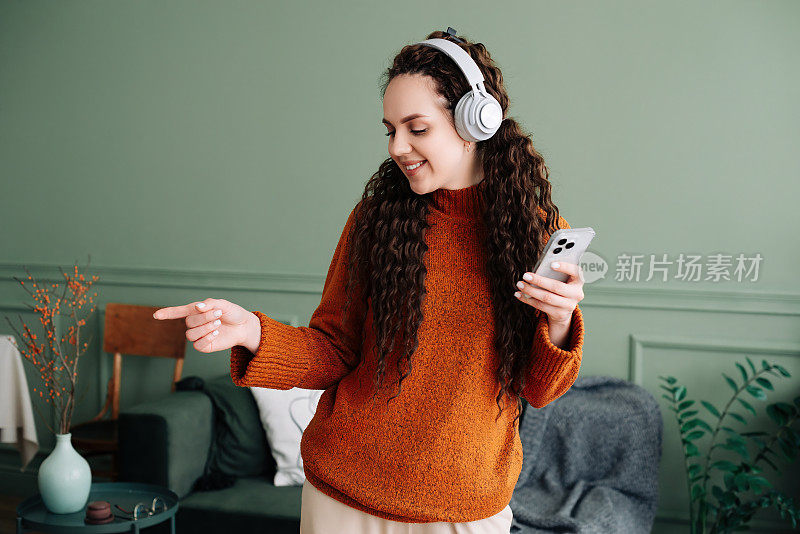 享受节奏:女人在客厅里戴着耳机在手机上随着音乐跳舞。戴着耳机用智能手机听音乐跳舞的快乐女人。