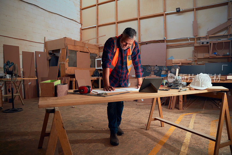 在木工工厂里，中年男子在数码平板电脑和桌子上的工具旁边工作