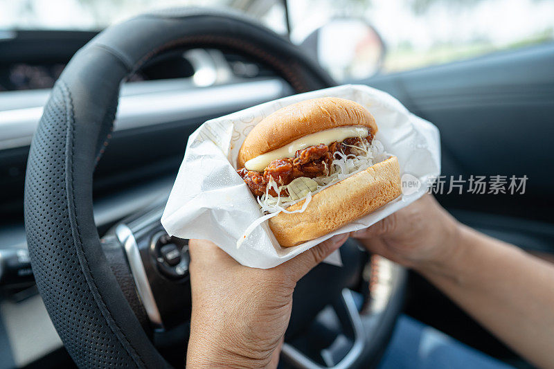 亚洲女士拿着汉堡在车里吃，危险而且有发生事故的危险。