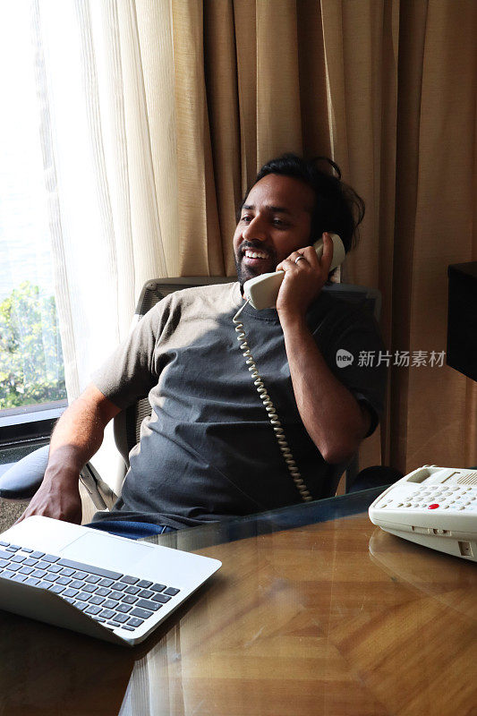印度人穿着休闲服在办公室工作，微笑着听座机电话，靠在办公椅上，把电话听筒放在耳边，笔记本电脑放在木制办公桌上，重点放在前景