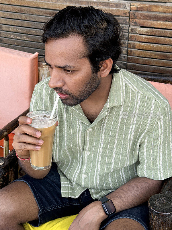 印度人坐在餐厅的露天用餐区，户外扶手椅，拿着一杯冰咖啡饮料，纸吸管，木板背景的面板墙，重点在前景
