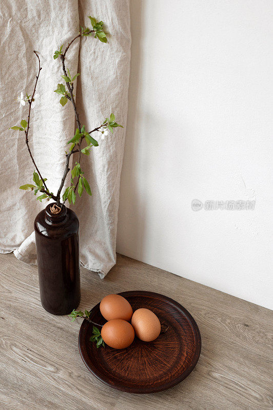 棕色陶瓷盘上的自然色彩蛋，餐桌上的绿色树枝花瓶，米色亚麻窗帘，空白墙，柔和的阳光阴影，优雅的可持续乡村复活节厨房内部