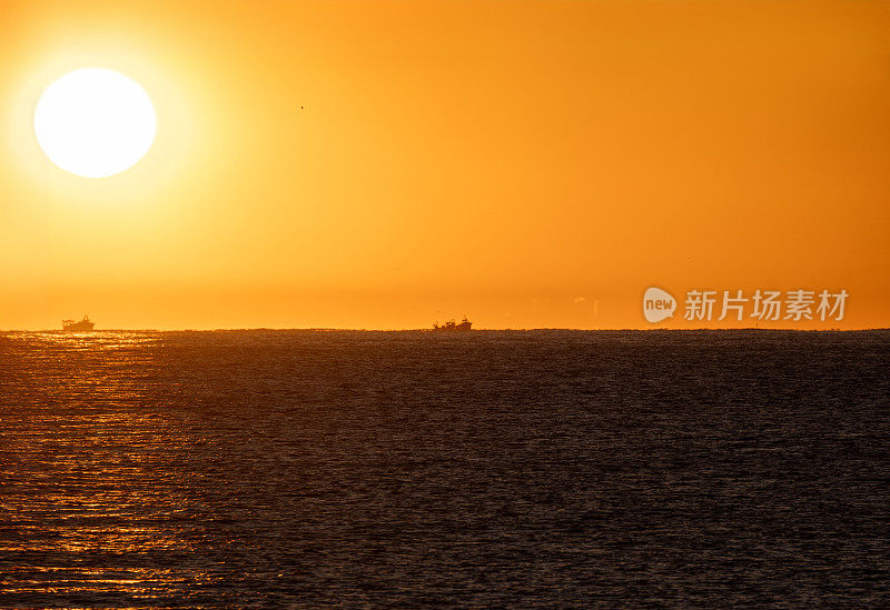 黄色的黎明太阳从复古的地中海地平线上出现，照亮了两艘渔船的剪影，在橙色的天空下出海捕虾。
