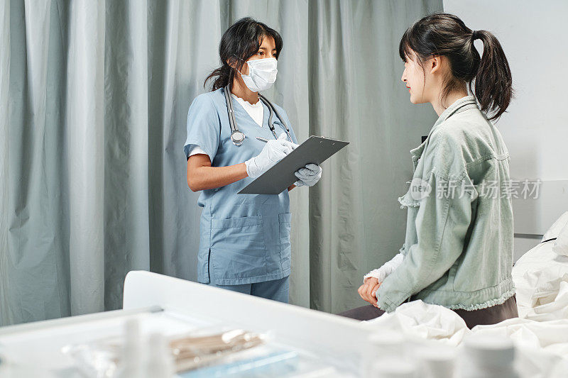 医生戴着口罩，拿着带医疗卡的剪贴板，在急诊室向年轻的亚裔女性提问