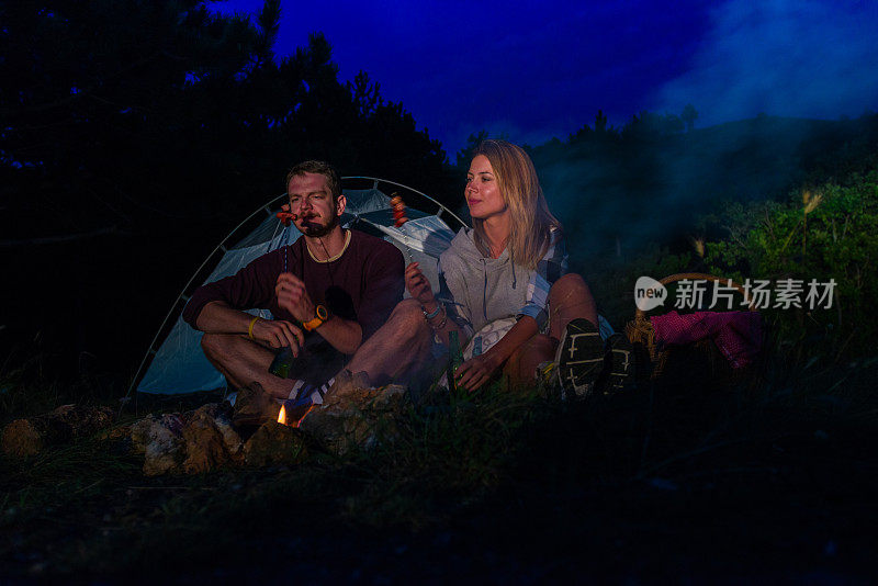 年轻的夫妇在篝火上烤香肠，在黄昏的森林山喝啤酒