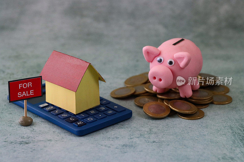纸板房子的形象站在计算器旁边出售的标志和小猪银行站在一堆硬币，灰色的背景，抵押贷款，家庭财务和储蓄的概念