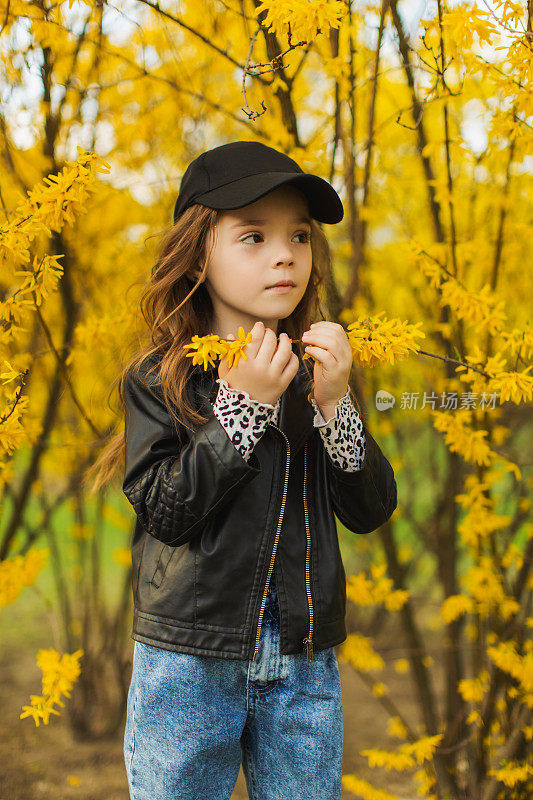 可爱的小女孩在大自然中有着长长的金色卷发。黄色开花灌木背景。亮片美妆。的童年。时尚的孩子。公主。时髦的衣服。闻到花朵。过敏