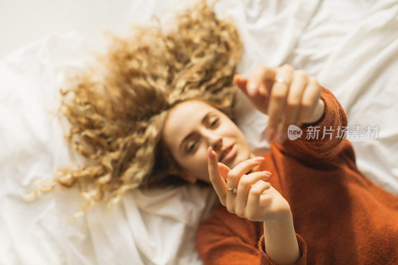 红头发卷发美丽的女孩躺在床上俯瞰。穿红毛衣的女人对着镜头举起手来。欢乐。白色背景上的长卷发。感觉幸福。开销