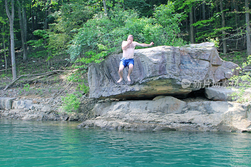 在美国西弗吉尼亚州泰加特湖州立公园的湖面上，一名男子从大岩石上跳入深蓝的湖中